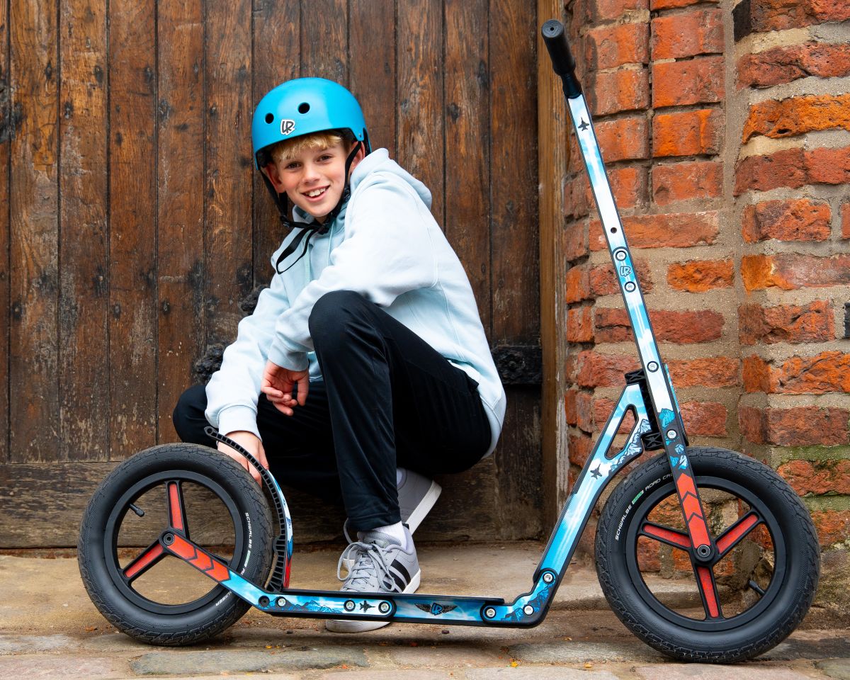 Child sitting on a LIFERYDER INFINITY big wheel scooter by a wooden gate and brick wall