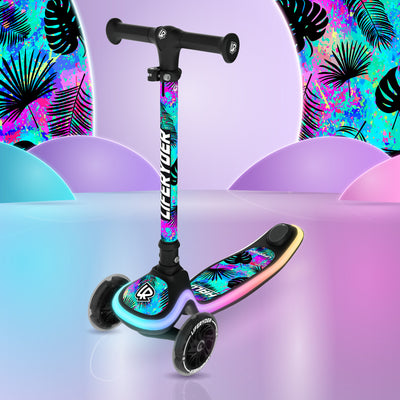 A children's three-wheel scooter with a black frame and colorful tropical leaf graphics, shown on a stylized pastel studio set with purple and blue lighting. 