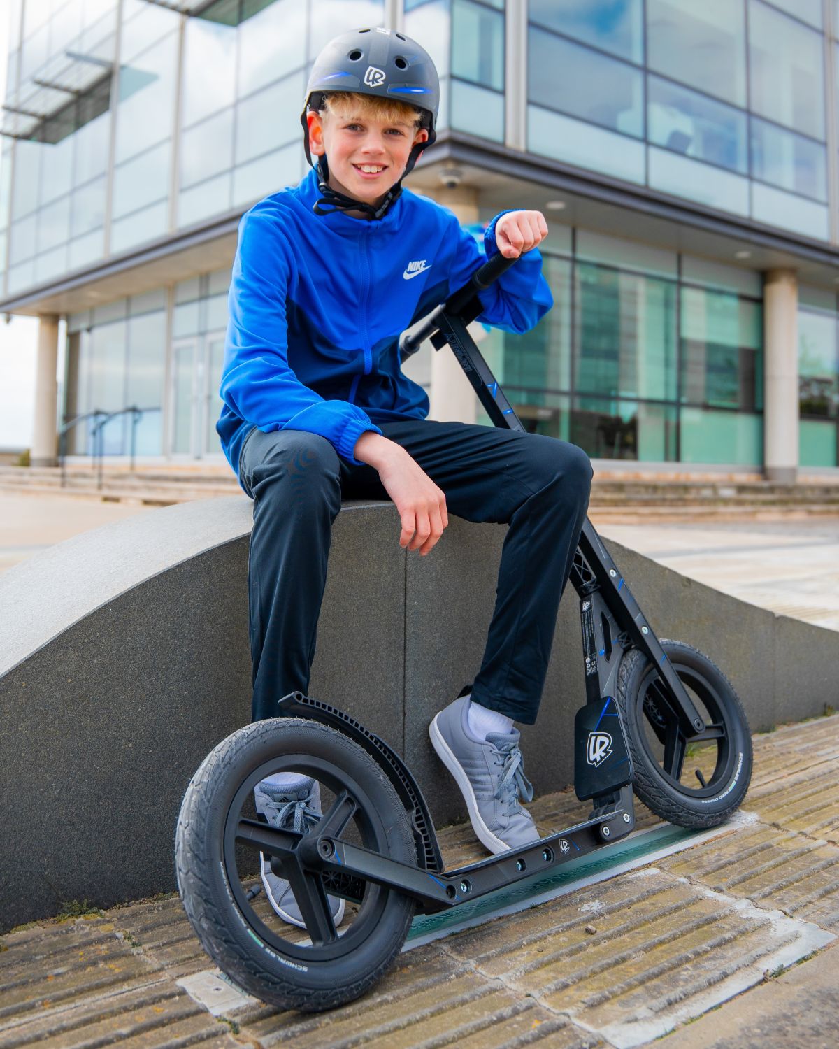 Child sitting on a LIFERYDER INFINITY big wheel scooter near a modern building